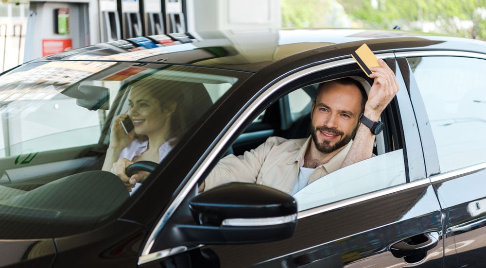 mulher e homem dentro de um carro em abastecimento. Na mão do homem está o Cartão Combustível Sem Parar Empresas, um cartão amarelo