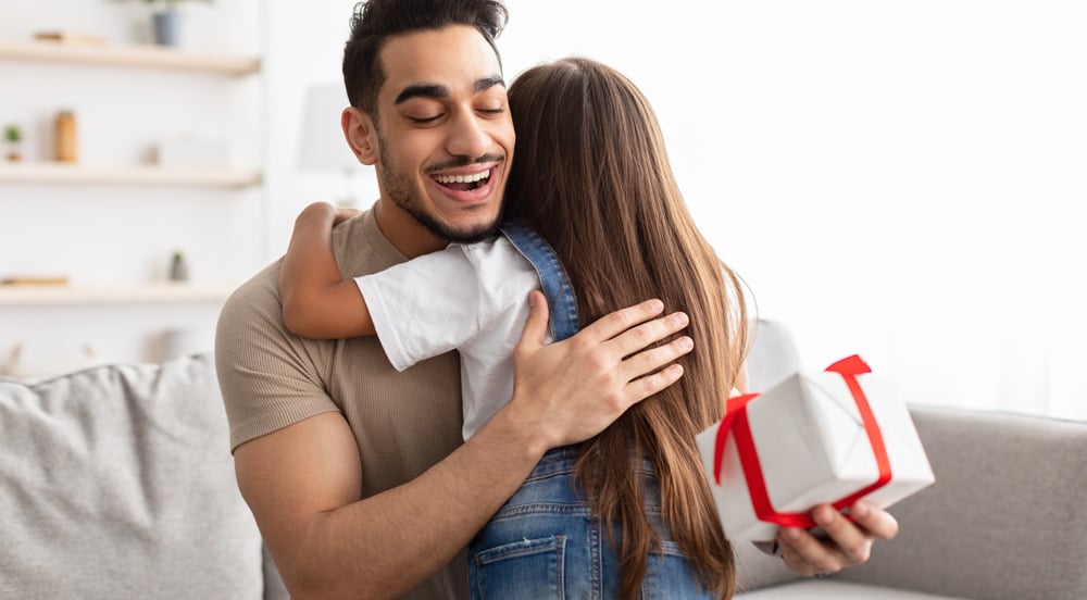 Homem sorrindo abraça sua filha. Em suas mãos tem uma caixinha de presente corporativo de dia dos pais que ganhou da empresa que usa o Cartão Presente da Sem Parar Empresas